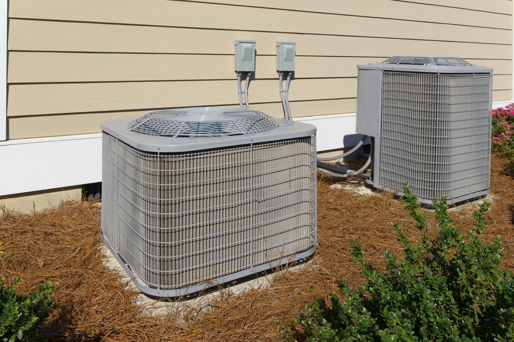 Two outdoor air conditioning units sit on concrete pads next to a beige house with mulch and bushes in the foreground.
