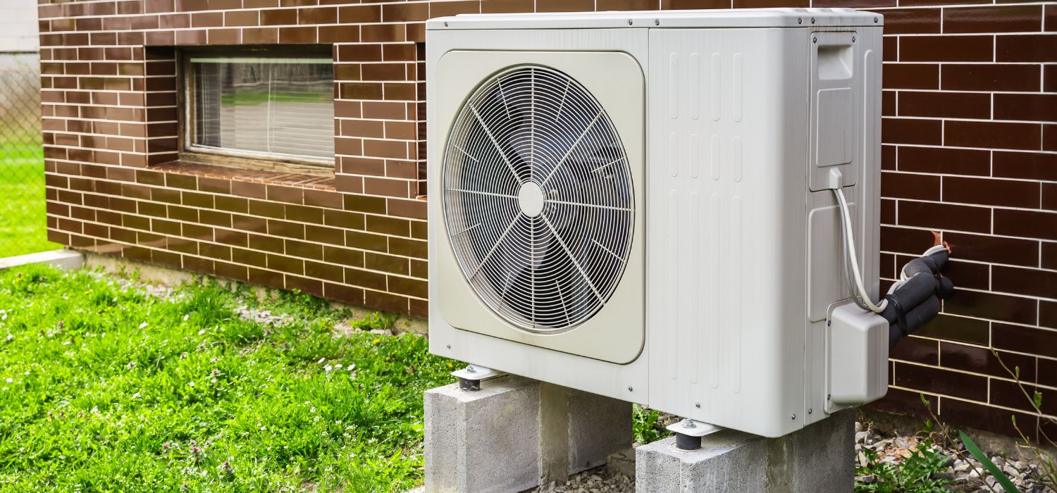 An outdoor air conditioning unit sits on concrete blocks next to a brown brick building, with grass and a window visible nearby.