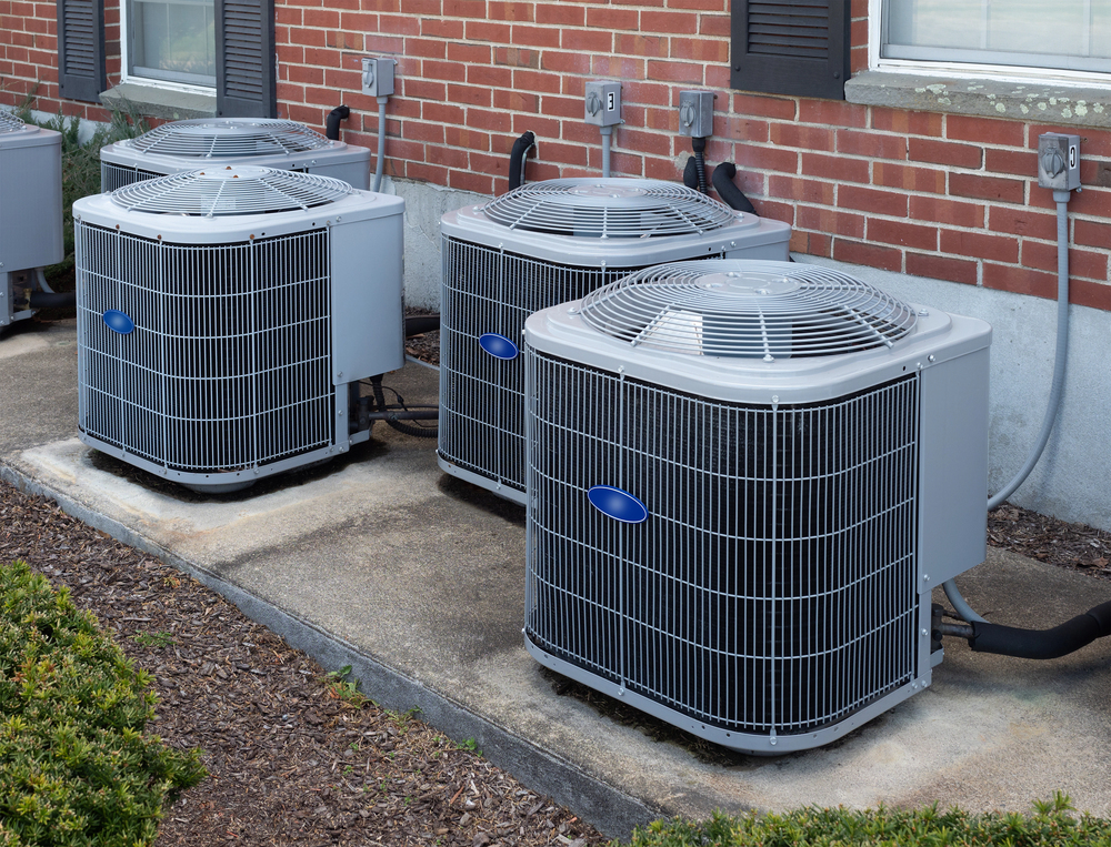 Three outdoor air conditioning units are positioned on a concrete pad beside a brick building, with electrical outlets and vents visible on the wall behind them.