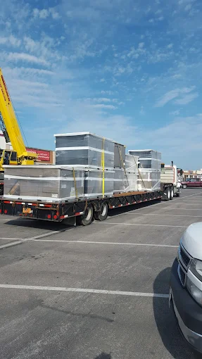 A flatbed truck loaded with large metal containers secured by straps is parked in a lot under a partly cloudy sky.