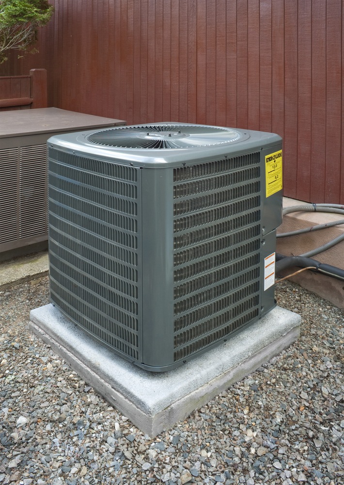 A gray outdoor air conditioning unit sits on a concrete pad surrounded by gravel, next to a wooden fence and another large unit.