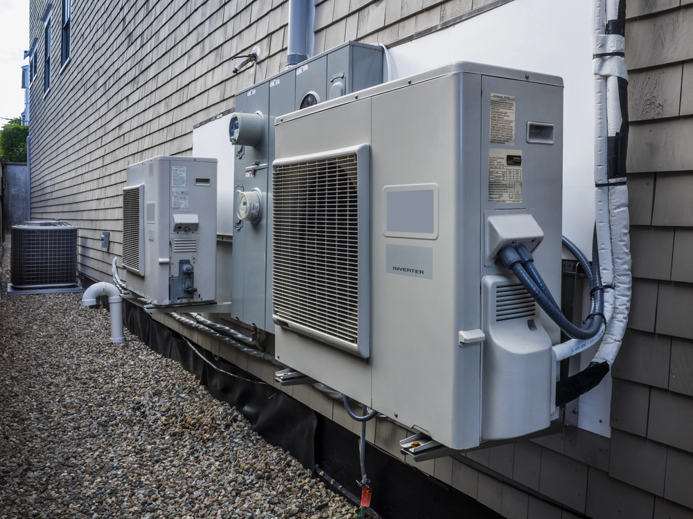 Several outdoor HVAC units mounted on the exterior wall of a building, with electrical and refrigerant lines attached, over a gravel ground.