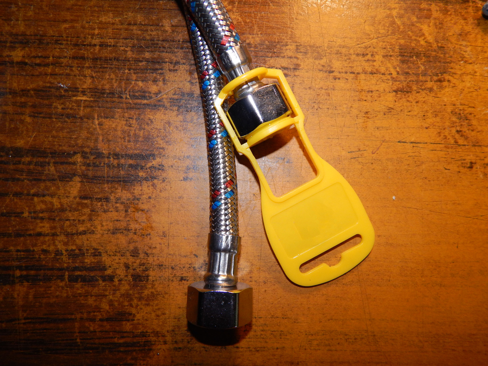 A braided metal hose with a hex nut fitting is next to a yellow plastic tag, all placed on a wooden surface.
