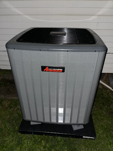 A gray Amana outdoor air conditioning unit sits on a concrete pad beside a white vinyl-sided building at night.