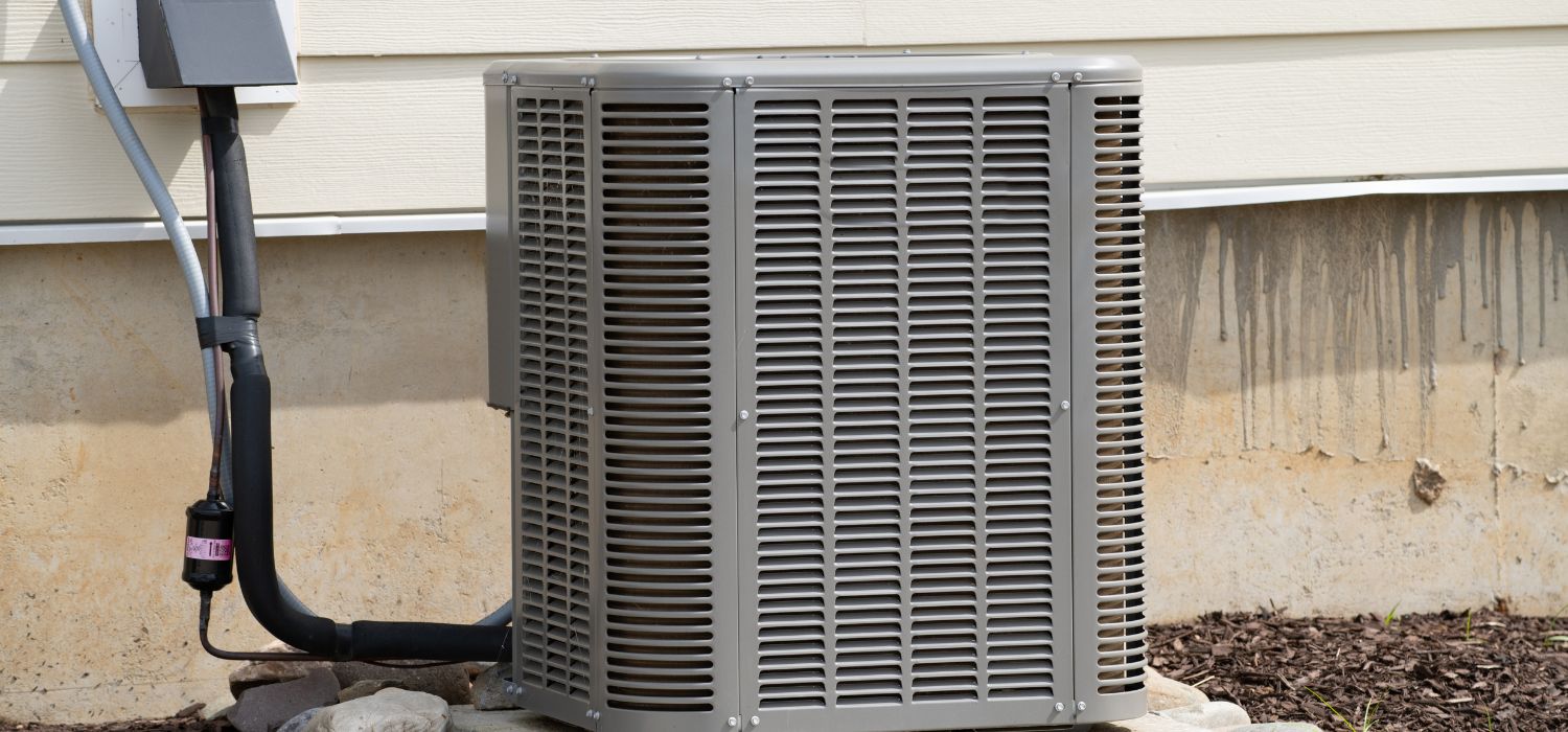 Outdoor central air conditioning unit installed next to a house exterior wall, featuring connected piping and electrical box—ideal for energy efficient heating in Mid-Missouri.