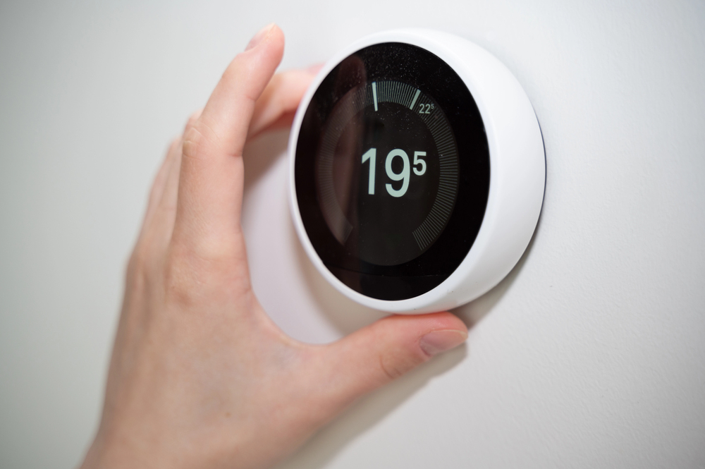 A hand adjusting a round digital thermostat mounted on a white wall, displaying a temperature setting of 19.5 degrees.