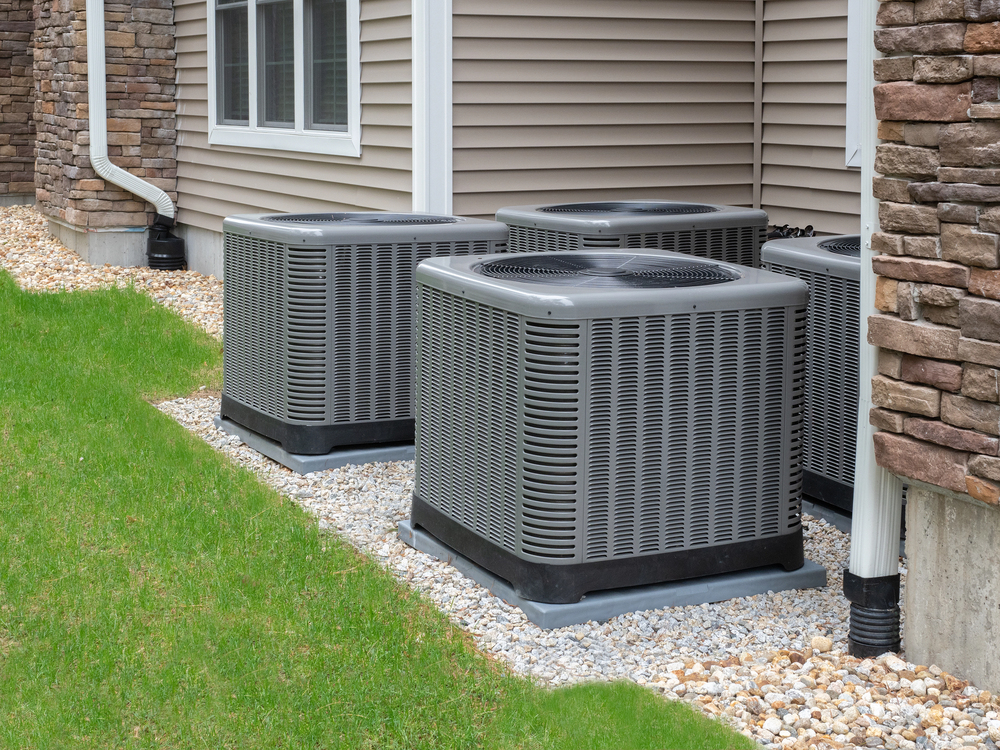 Three outdoor air conditioning units from a recent AC installation are placed on gravel beds next to the exterior wall of a house with siding and stone accents.